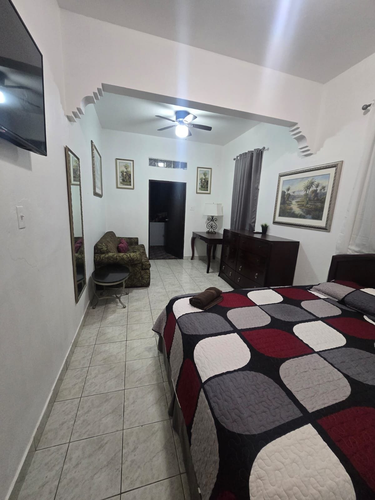 Un dormitorio con cama, escritorio, silla, ventilador de techo y televisor montado en la pared.