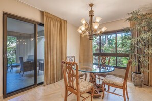 A dining area with a glass table and wooden chairs, a chandelier, and a view of the outdoors through large windows. - 160 Steps from kitchen sink to sand! Platinum Interior. 1431sf - New Ownership! (Lahaina)