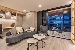 Un salon moderne avec un canapé gris aux lignes courbes, une table basse ronde et un fauteuil gris. On aperçoit un balcon avec des meubles d’extérieur et, au loin, un immeuble.