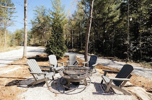 A circular fire pit surrounded by four Adirondack chairs in a forested area.