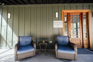 A covered porch with two wicker chairs and a small table, a wooden door with glass panels, and a sign indicating the establishment year.