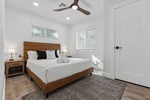 Une chambre à coucher comprenant un lit, une table de chevet, une lampe, un ventilateur de plafond et deux fenêtres équipées de stores.