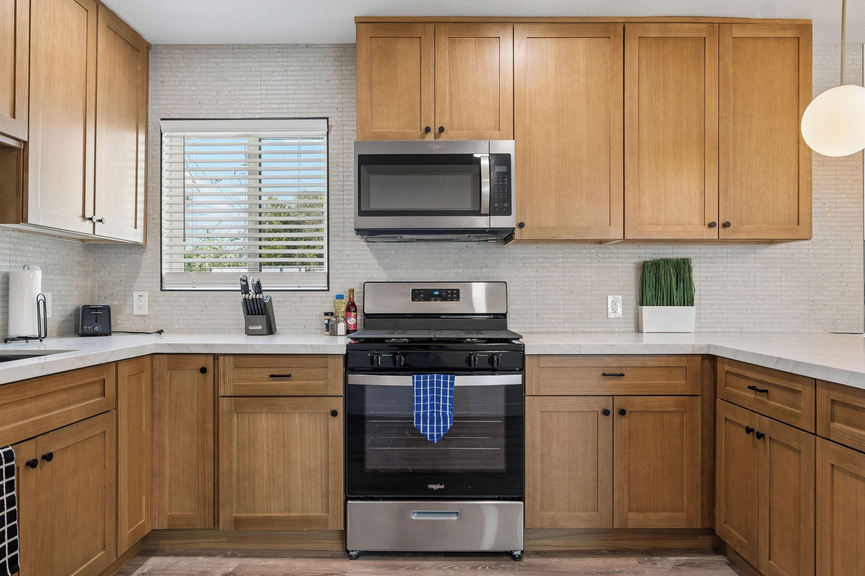 A modern kitchen with wooden cabinets, a stainless steel stove, a microwave, and a window with blinds.