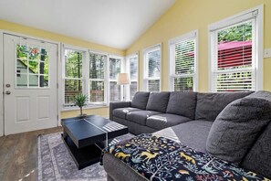 A living room with a gray sectional sofa, a black coffee table, and a patterned ottoman. There are white-framed windows with blinds, and a door with a glass panel. - Arrowhead Hideaway Hot Tub Fire Pit Game Room (Monroe County)
