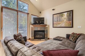A living room with a stone fireplace, a large flat-screen TV, and a large window with blinds. - Beautiful 3 Bedroom + Den Townhome in Breckenridge, Colorado (Breckenridge)