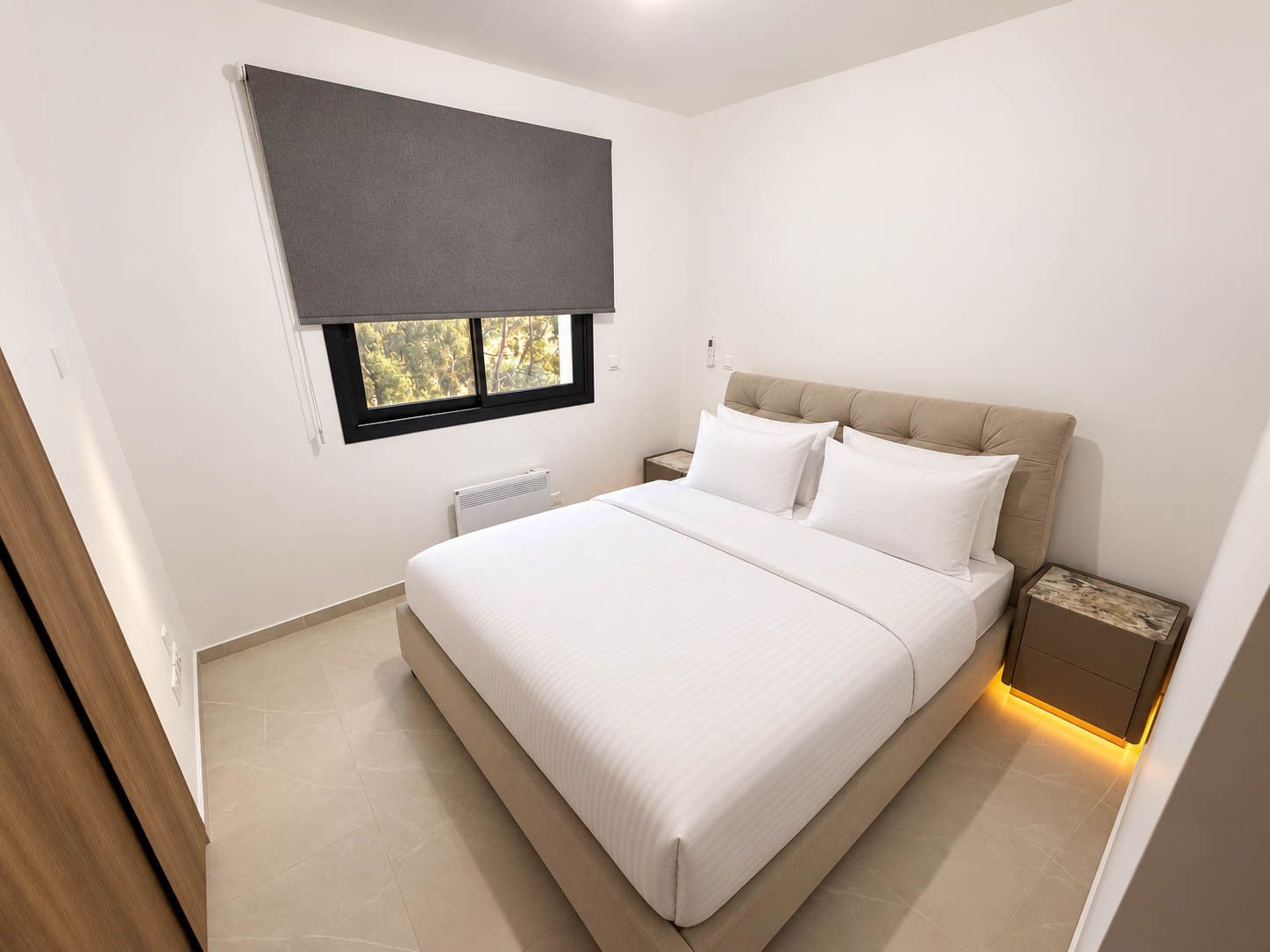 A modern bedroom with a large bed, a window with a blind, a bedside table, and a radiator.