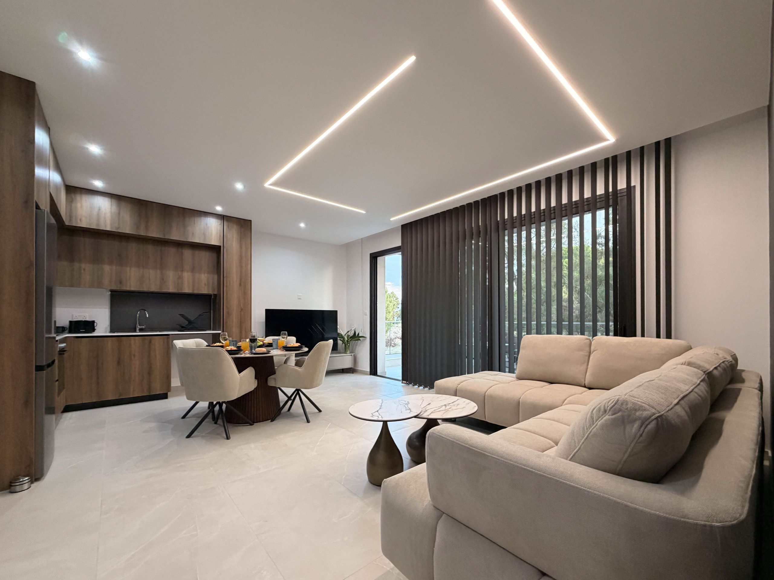 A modern living room with a sofa, a dining area with a table and chairs, and a kitchen with wooden cabinets.