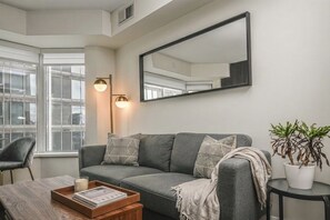 A modern living room with a grey sofa, a coffee table with a magazine, a floor lamp, and a potted plant. - Radiant 1BR Condo Yorkville (Toronto)