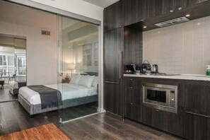 A modern hotel room with a glass partition separating the bedroom from the living area, a kitchenette with a microwave, and a view of the cityscape. - Radiant 1BR Condo Yorkville (Toronto)