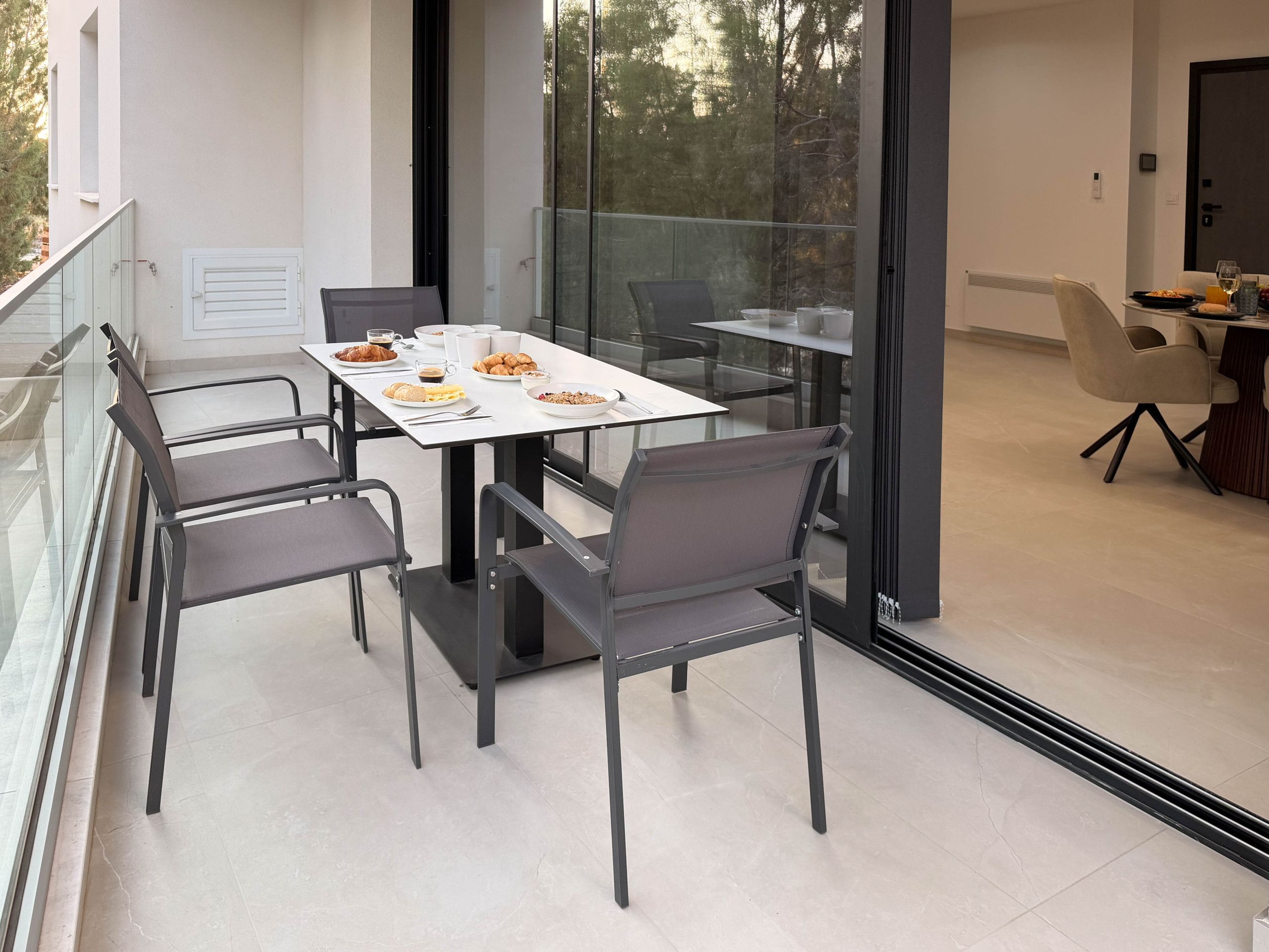 A modern dining area with a glass table and chairs, a balcony with a railing, and a view of trees.