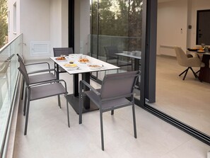 A modern dining area with a glass table and chairs, a balcony with a railing, and a view of trees.