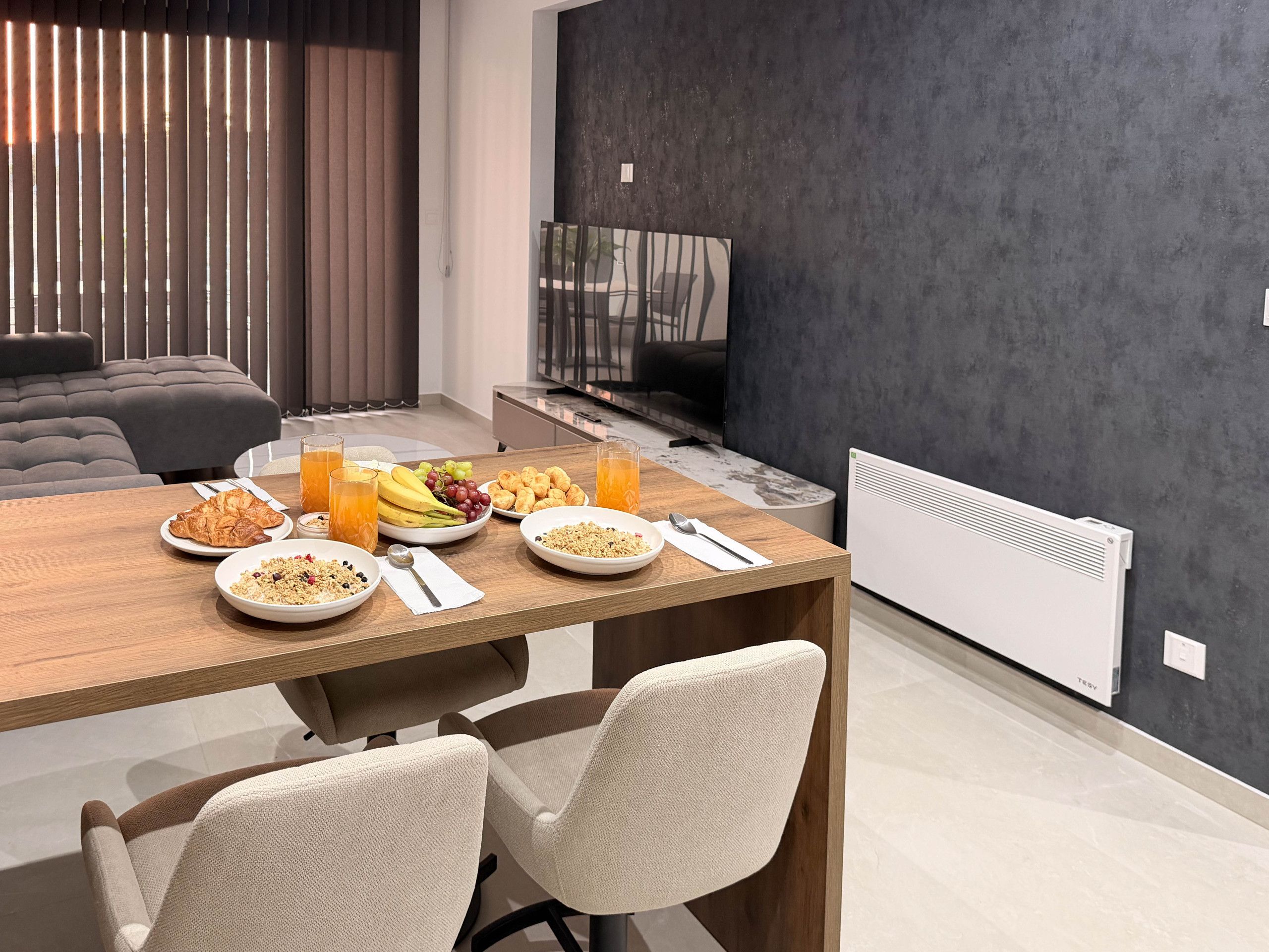 A modern dining area with a wooden table set for breakfast, including plates of cereal, fruit, and pastries.