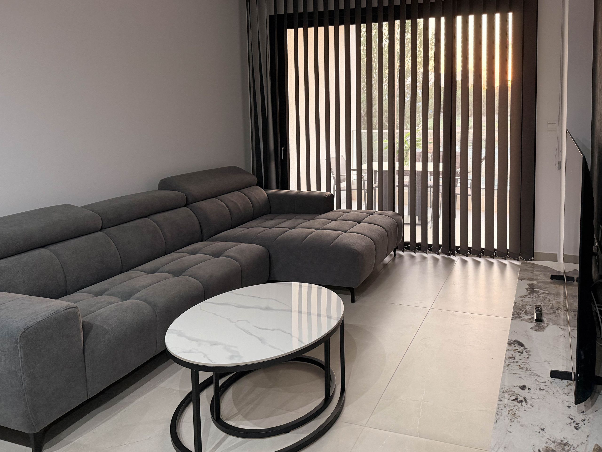 A modern living room with a grey sectional sofa, a round coffee table, and a large window with vertical blinds.