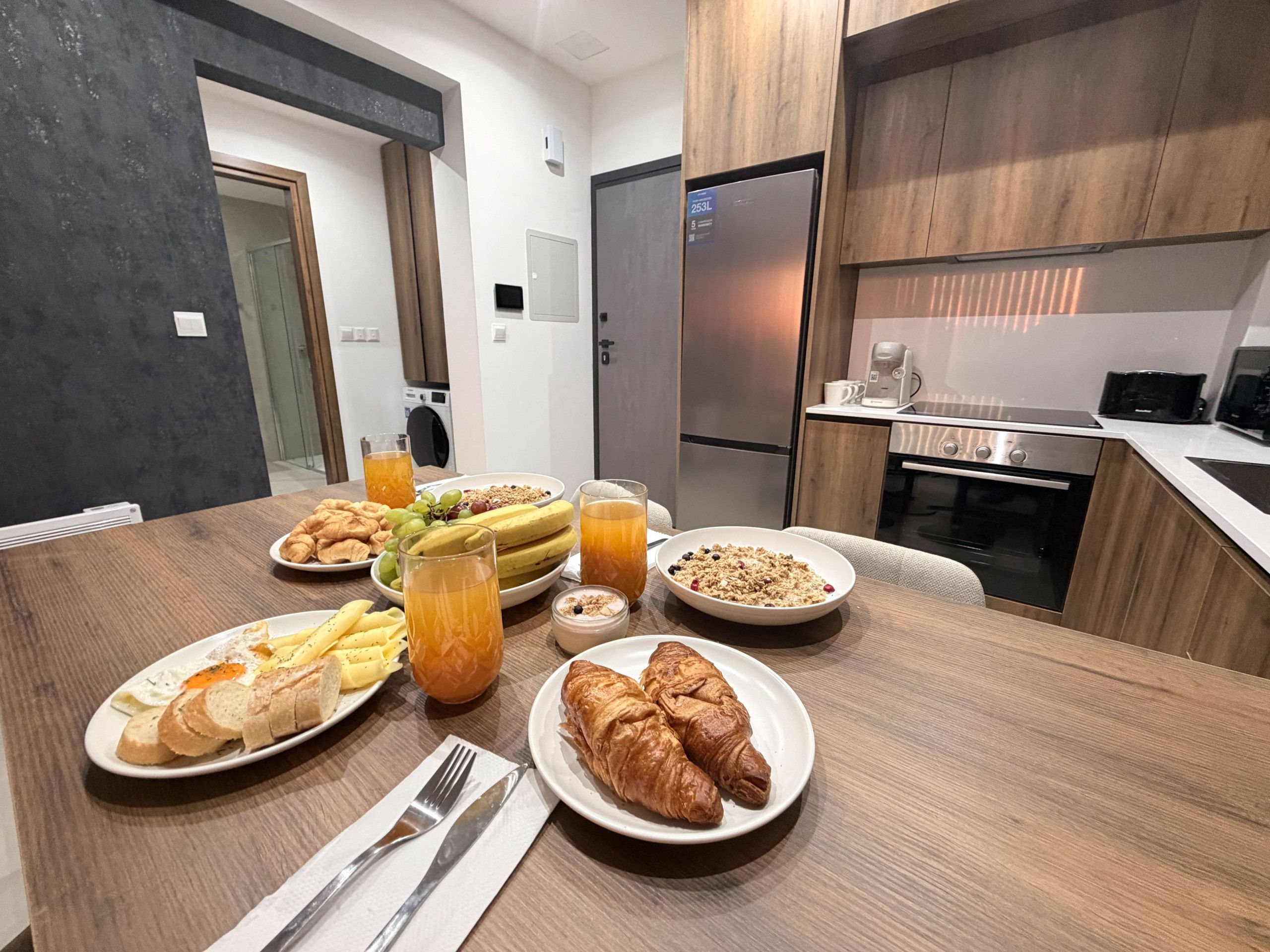 A breakfast setting with croissants, fruit, cereal, and juice on a wooden table in a modern kitchen.