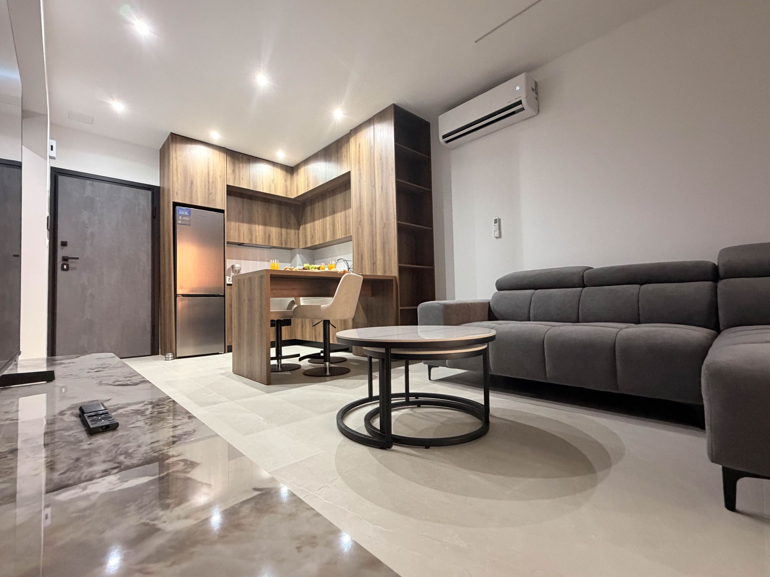 A modern living room with a grey sofa, a round coffee table, and a kitchen area with wooden cabinets and a built-in fridge.