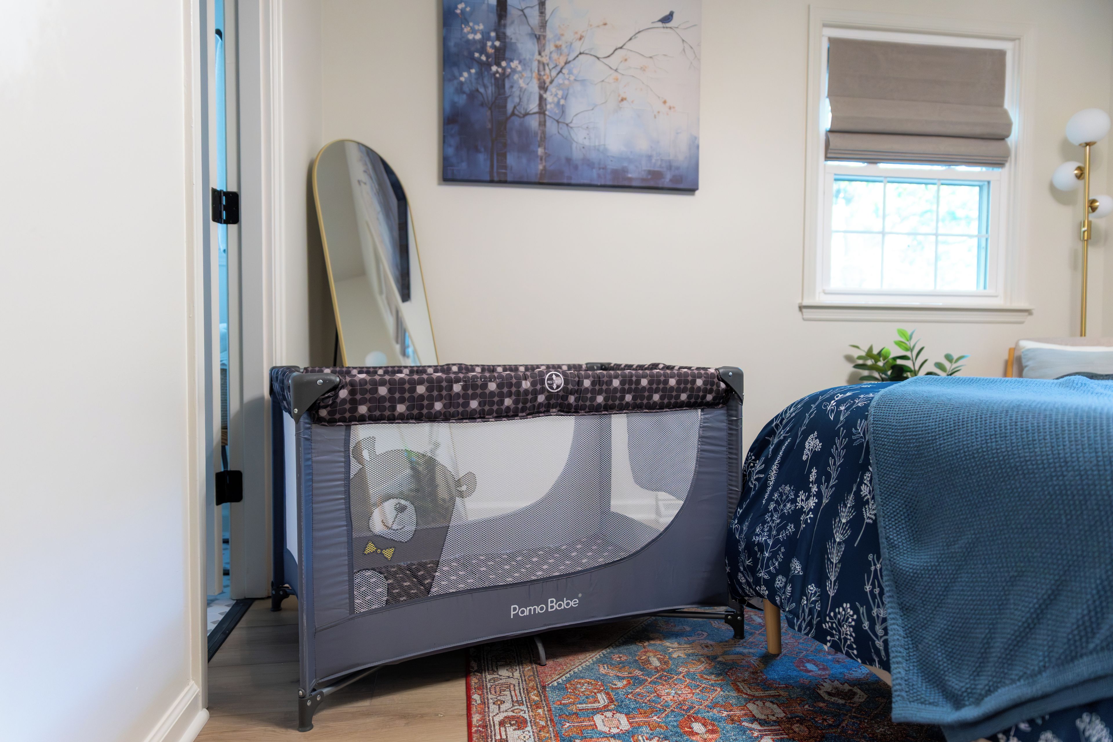 A room with a crib, a window with blinds, a painting on the wall, and a patterned rug.
