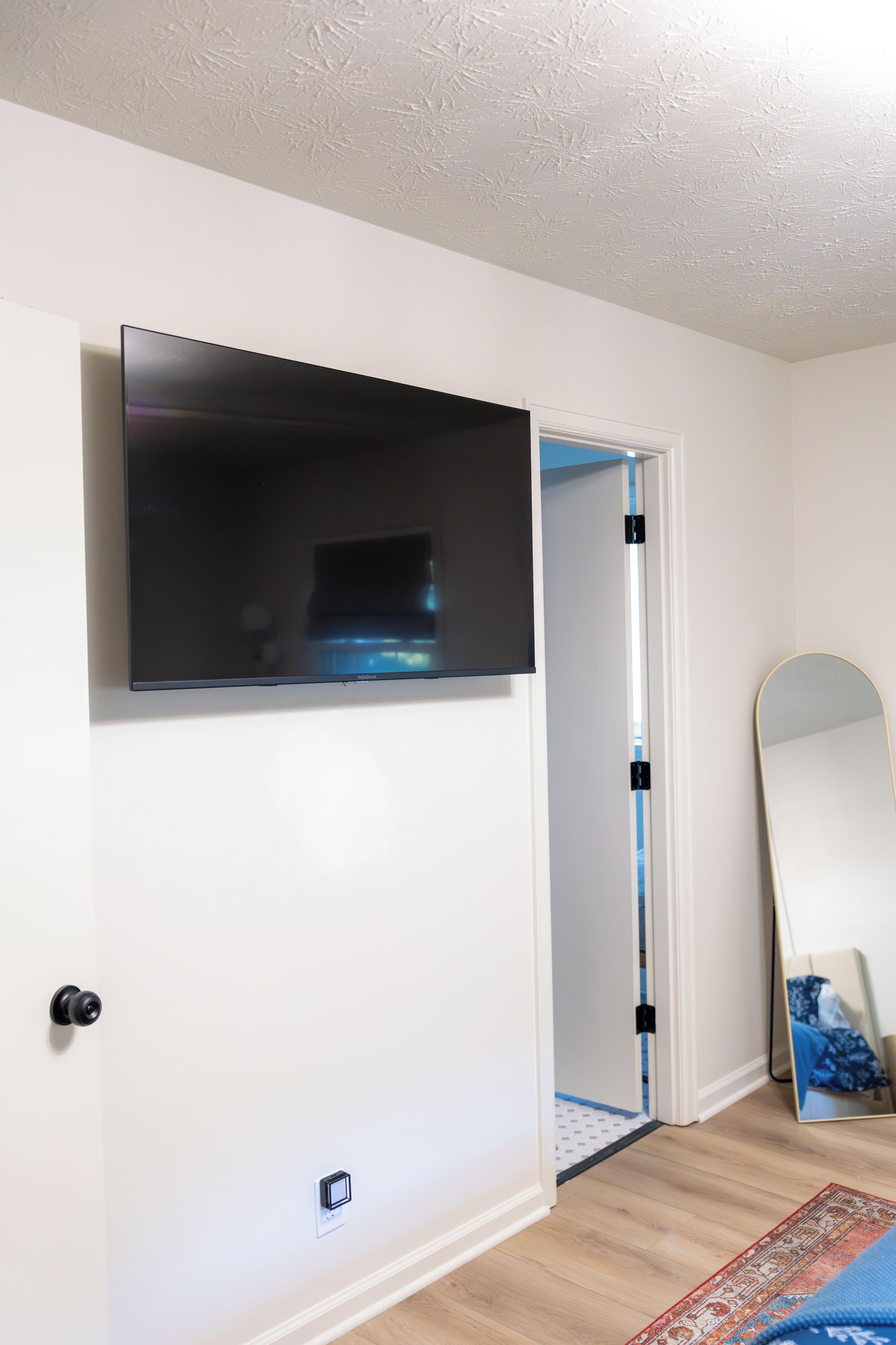A room with a flat-screen TV mounted on the wall, a door with a mirror, and a patterned rug on the floor.