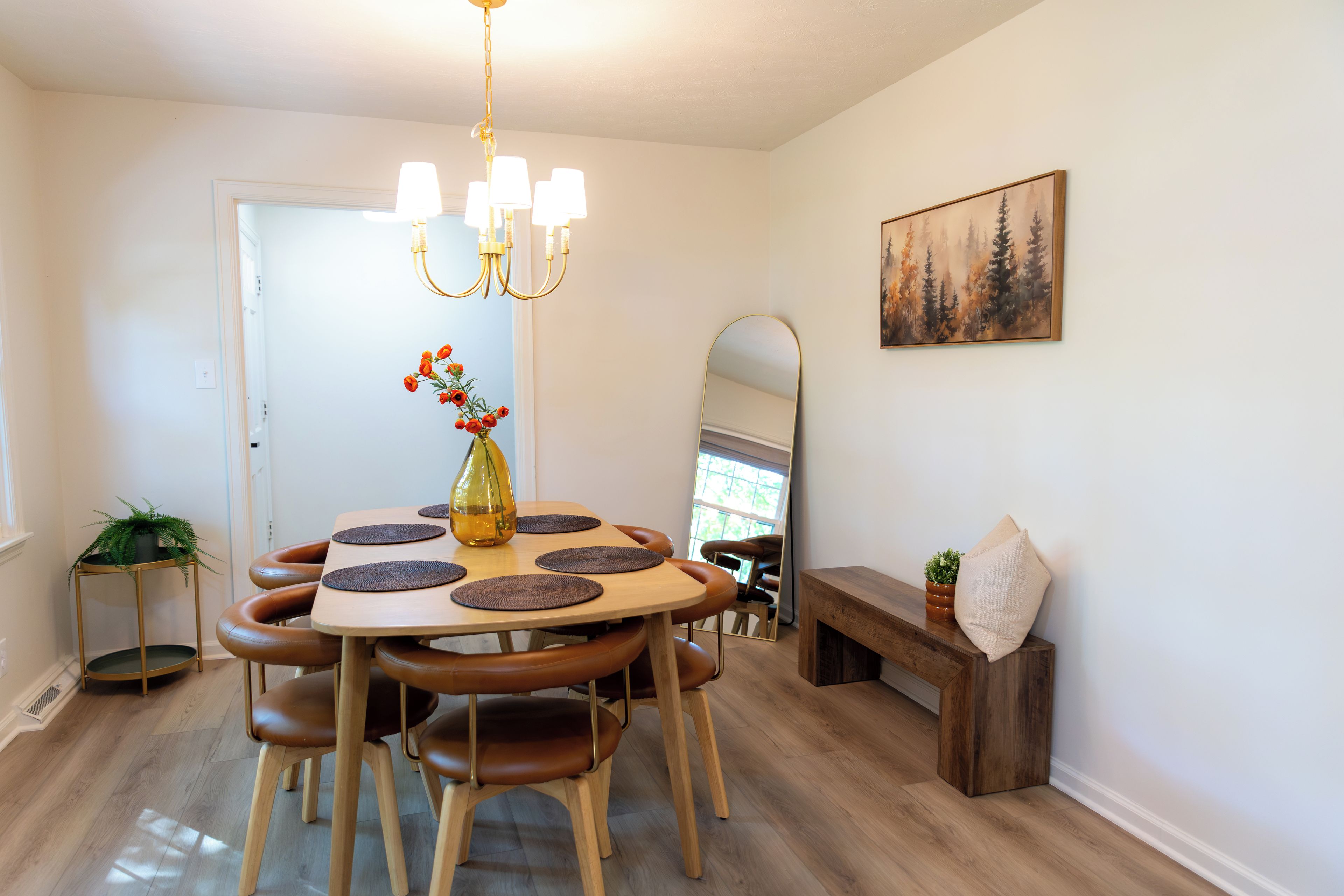 Spiseområde med et rundt bord, stole, en lysekrone, en vase med blomster, et spejl og et billede på væggen.
