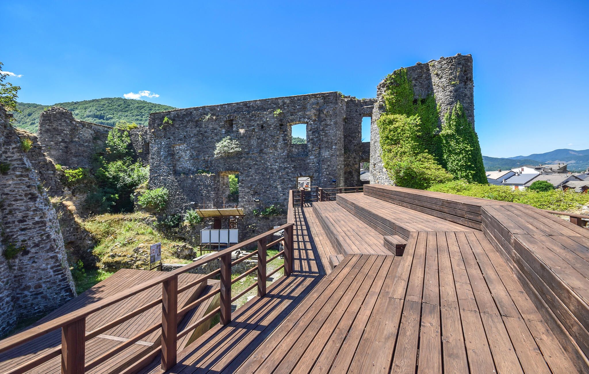 A wooden deck leading to a stone structure with a view of a mountain and houses in the distance.