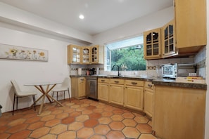 A kitchen with wooden cabinets, a dining table with chairs, a window with a view of greenery, and a tiled floor.