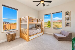 Une chambre avec un lit superposé, un ventilateur de plafond, de grandes fenêtres donnant sur un paysage désertique, un fauteuil vert et un panier en osier.