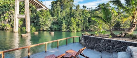 A concrete deck with lounge chairs and a table overlooking a river, with a bridge in the background.