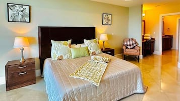 A well-lit bedroom with a large bed, two bedside tables with lamps, a chair, and artwork on the walls.