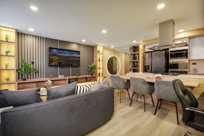 Una sala moderna con un sofá gris, un televisor montado en la pared y una zona de cocina con estufa y armarios.
