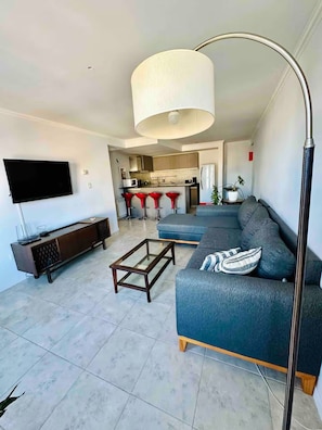 A modern living room with a sofa, a coffee table, a TV, and a kitchen in the background. - Hermoso departamento céntrico con cochera (San Carlos de Bariloche)
