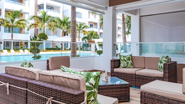 Un área de descanso moderna al aire libre con mobiliario de mimbre, una mesa de centro de vidrio y vistas a una piscina y palmeras.