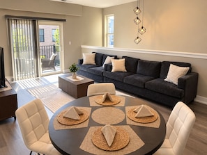 A modern living room with a dark grey sofa, a round dining table with placemats, and a television on a wooden stand. - 2-bedroom furnished luxury apartment Huntley Crossing (Huntley)