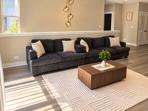 Ein modernes Wohnzimmer mit einem dunkelgrauen Ecksofa, einem Holztisch und einem gemusterten Teppich.