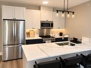 A modern kitchen with stainless steel appliances, a marble countertop, and black bar stools. - 2-bedroom furnished luxury apartment Huntley Crossing (Huntley)
