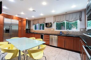 Una cocina moderna con armarios de madera, electrodomésticos de acero inoxidable, una mesa de comedor de vidrio con sillas amarillas y una ventana con cortinas.