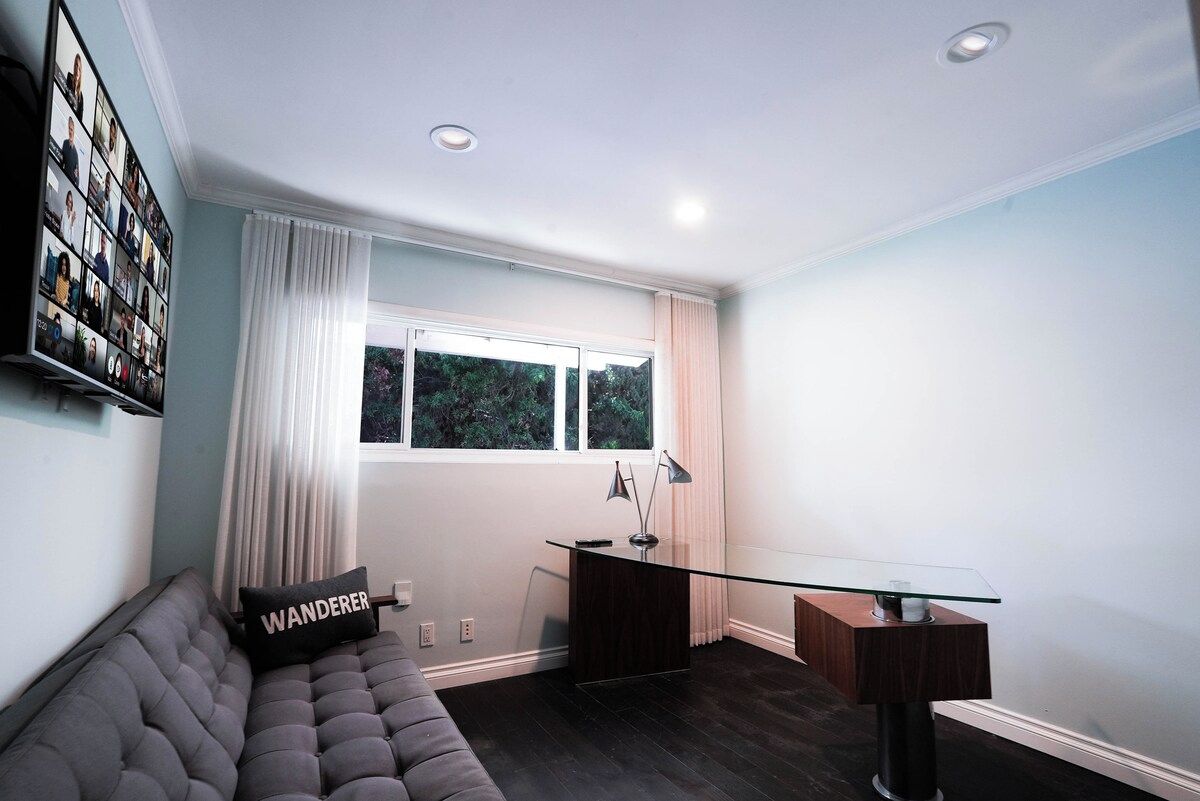 Una sala de estar moderna con un escritorio de vidrio, un sofá acolchado y un amplio ventanal con cortinas.
