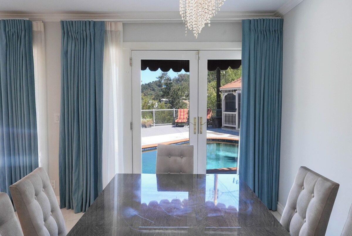 A dining area with a glass table, chairs, and a view of a swimming pool and outdoor seating.