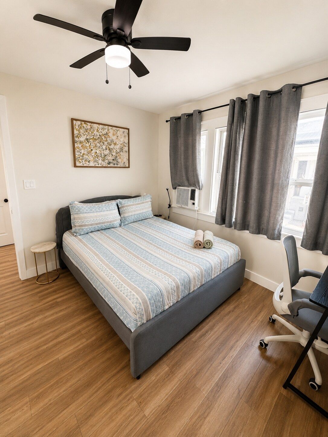 Un dormitorio con cama, ventilador de techo, ventana con cortinas, una mesita redonda y una silla.