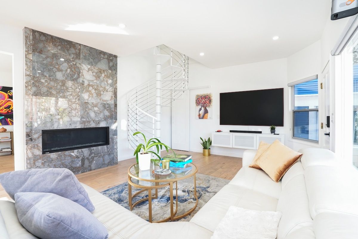 Una sala de estar moderna con pared de mármol, una escalera espiral, un televisor de pantalla plana, un sofá blanco y una mesa de centro redonda.