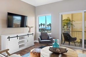 A modern living room with a flat-screen TV, a white console with drawers, a grey armchair, a round coffee table with decorative items, and a view of palm trees through a large window. - The Castle Tower Neblina (Oceanside)