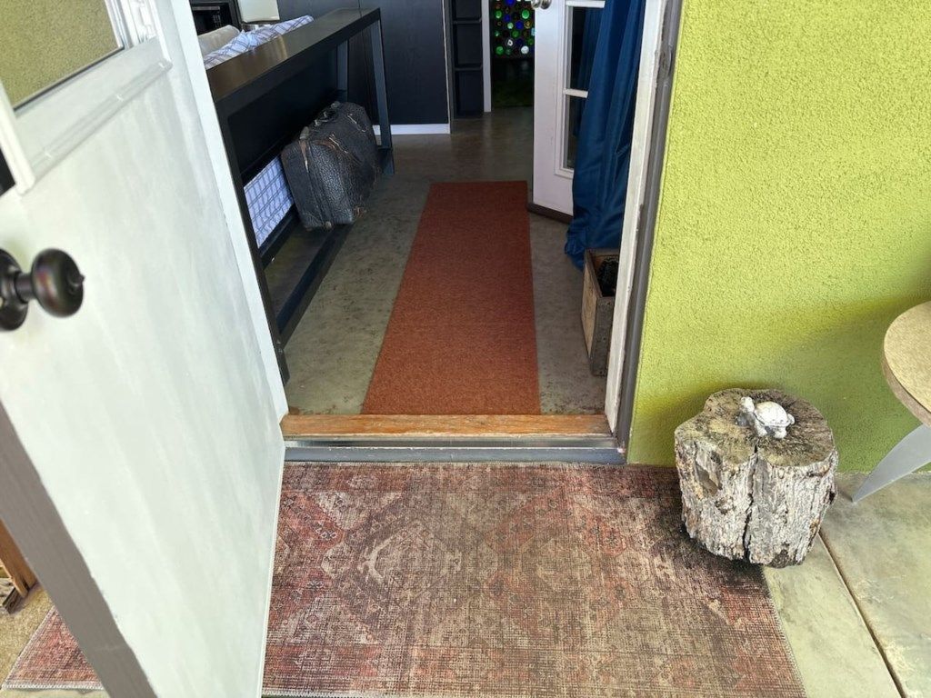En hall med ett brunt golv som leder in till ett rum, en träbänk med väskor på och en liten trädstump.