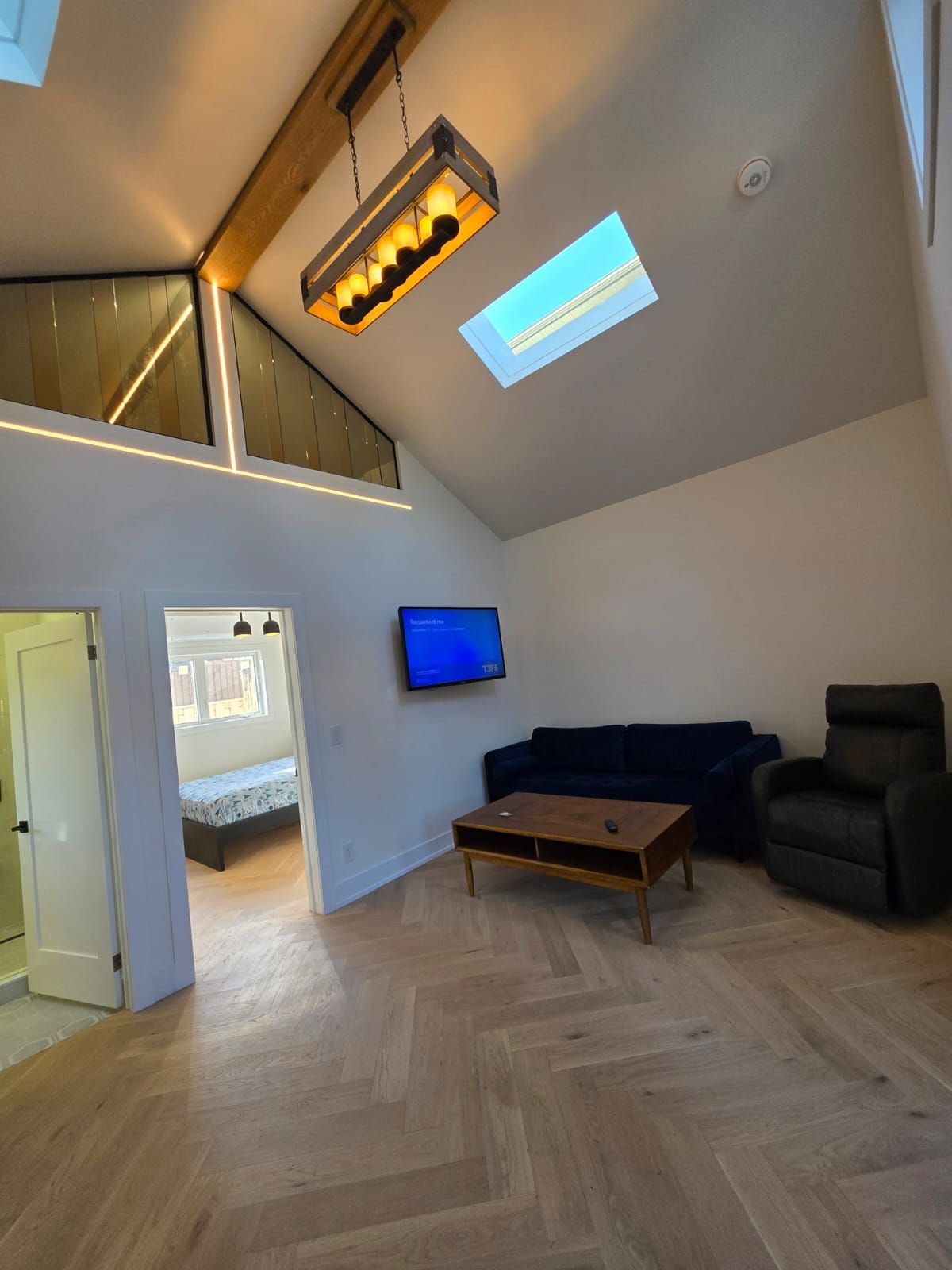 A spacious living area with a high ceiling, a television mounted on the wall, a dark blue sofa, a wooden coffee table, and a staircase leading to an upper level.