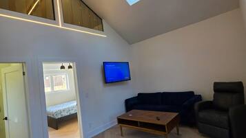 A spacious living area with a high ceiling, a television mounted on the wall, a dark blue sofa, a wooden coffee table, and a staircase leading to an upper level.