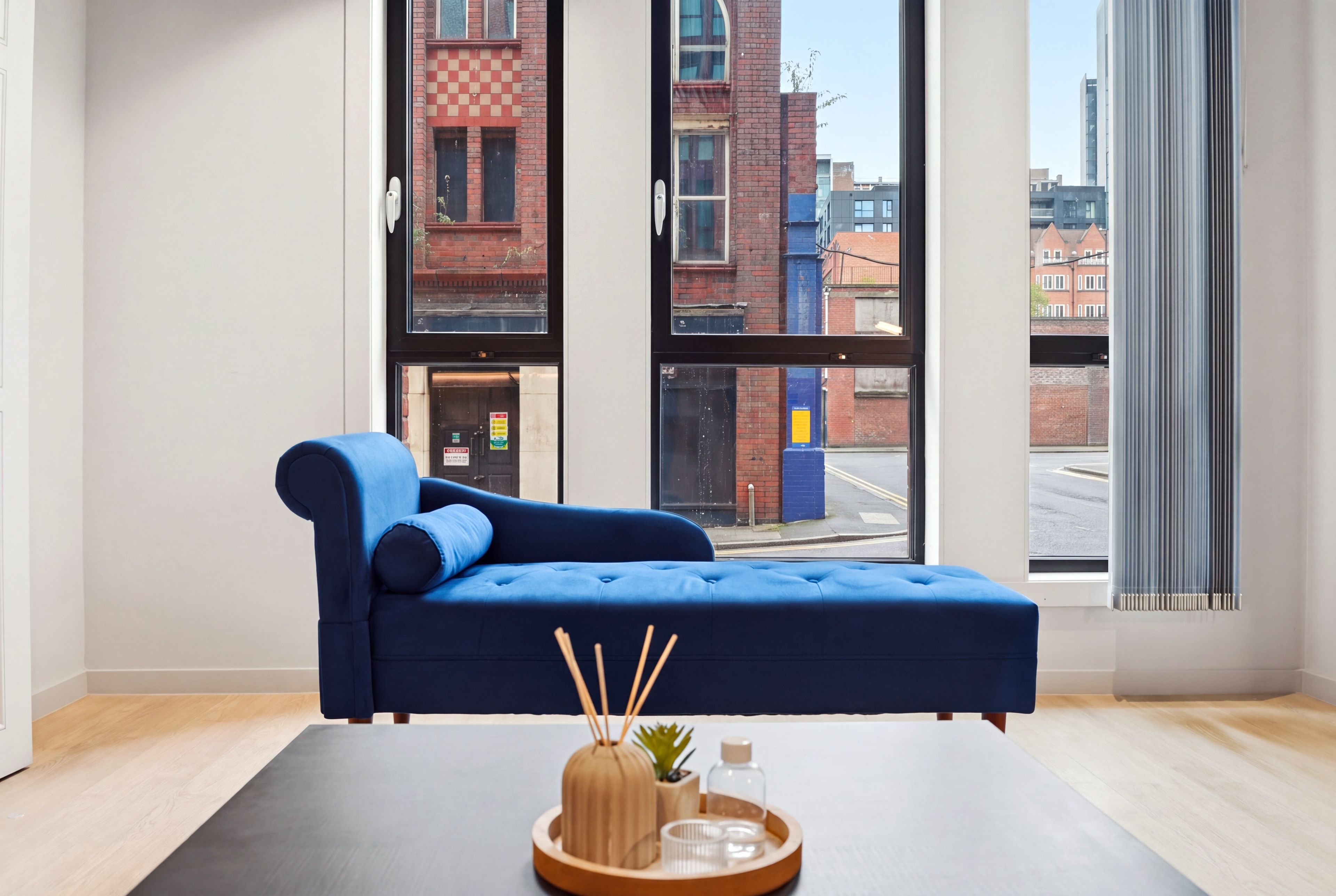 Une chambre moderne avec un canapé bleu, une petite table sur laquelle est posée une plante, et de grandes fenêtres offrant une vue sur la ville.