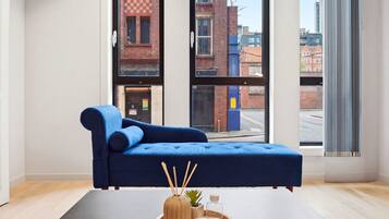 Une chambre moderne avec un canapé bleu, une petite table sur laquelle est posée une plante, et de grandes fenêtres offrant une vue sur la ville.