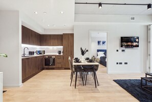 Eine moderne Küche mit Holzschränken, ein Essbereich mit Tisch und Stühlen sowie ein Wohnzimmer mit an der Wand befestigtem Fernseher.