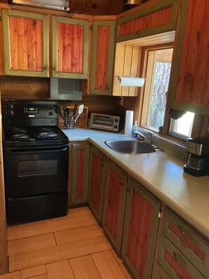 A kitchen with wooden cabinets, a stove, a toaster, and a sink. - Cute little cabin in the middle of the woods, yet still right in town. (Beaverdell)