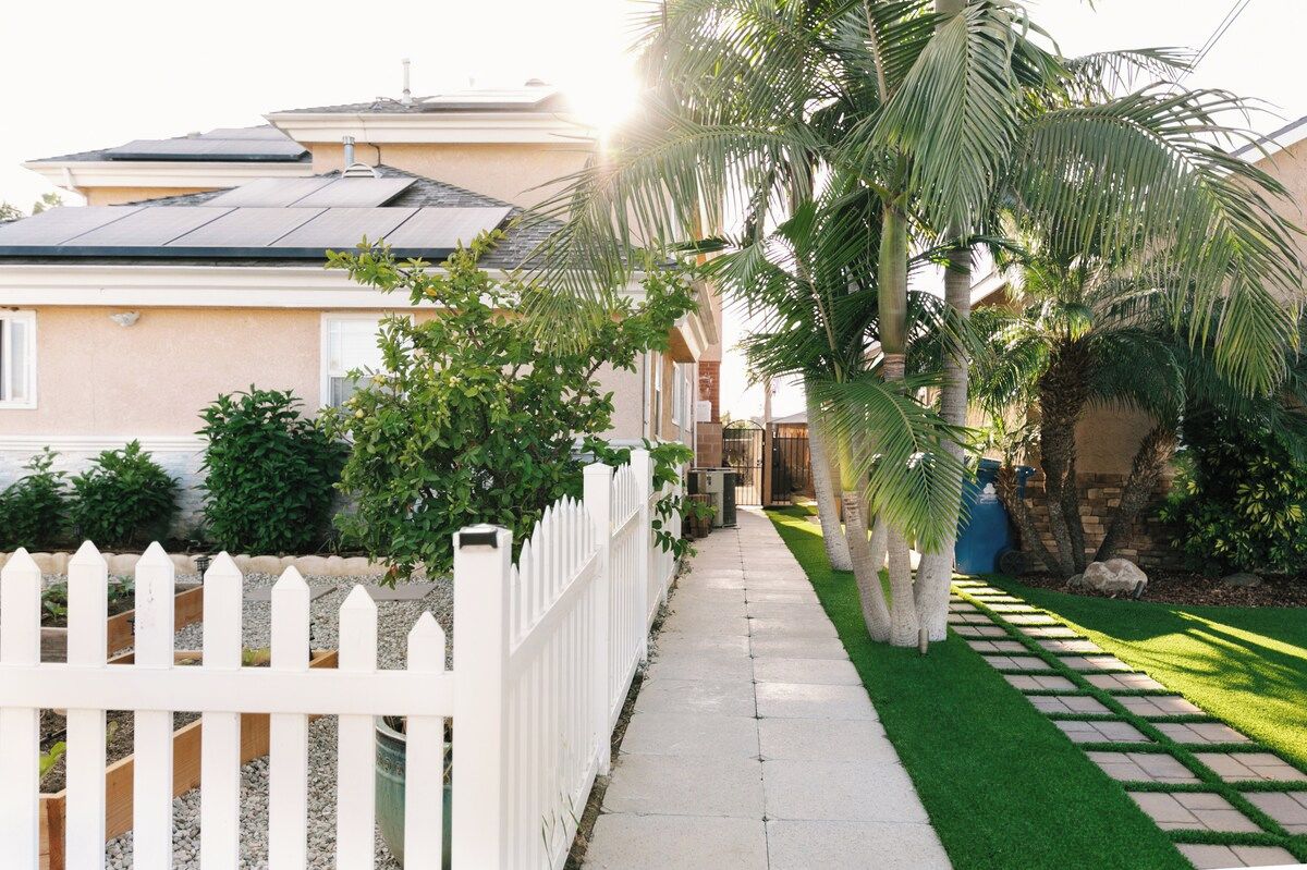 Una propiedad residencial con entrada pavimentada, cerca blanca en forma de piquete y diversa vegetación, incluyendo palmeras y arbustos.
