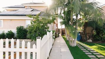 Una propiedad residencial con entrada pavimentada, cerca blanca en forma de piquete y diversa vegetación, incluyendo palmeras y arbustos.