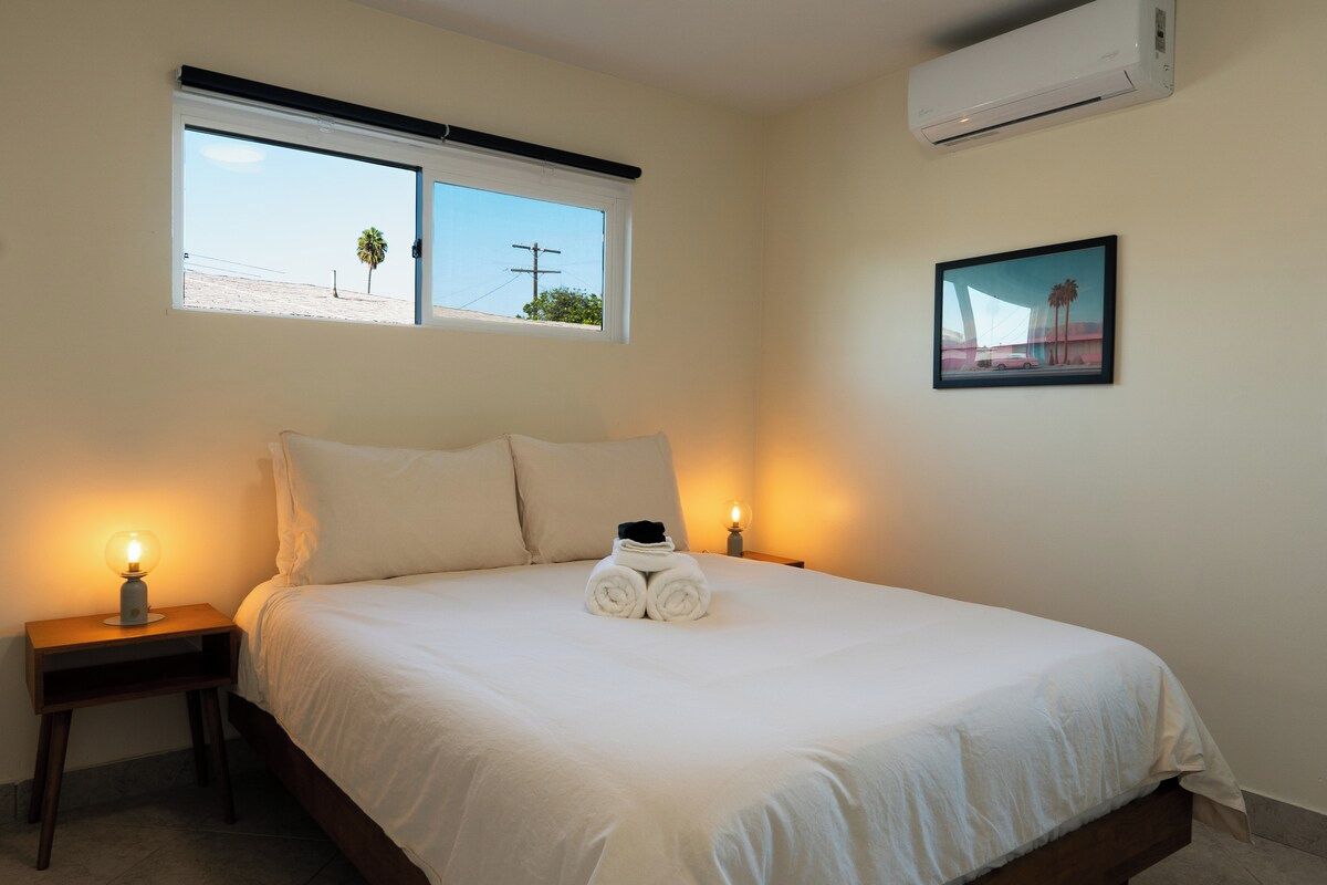 A bedroom with a bed, two bedside lamps, a window with a view, and an air conditioner.
