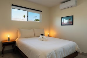 A bedroom with a bed, two bedside lamps, a window with a view, and an air conditioner.